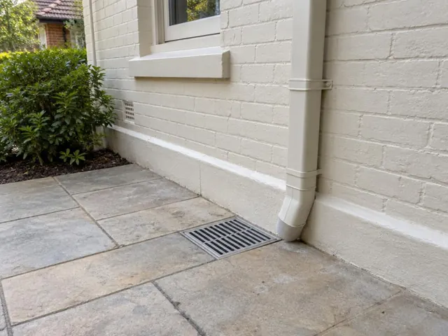 Outdoor gully grate beside a cream brick wall and downpipe on paved ground in Gordon