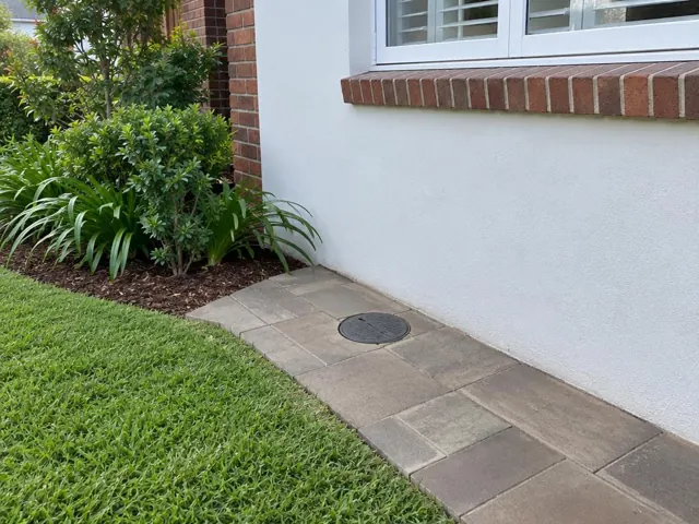 Flush circular access cover set in reinstated paving beside a brick and rendered house wall in Wahroonga