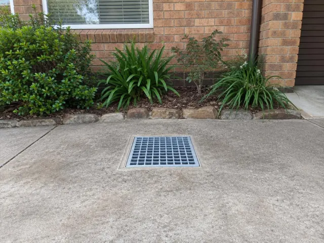 Flush square stormwater grate in a dry concrete driveway beside brick garden beds in St Ives